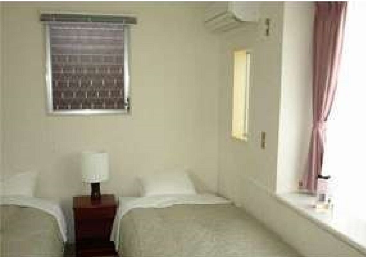 A hotel room with two beds, a nightstand with a lamp, a window with curtains, and an air conditioning unit.