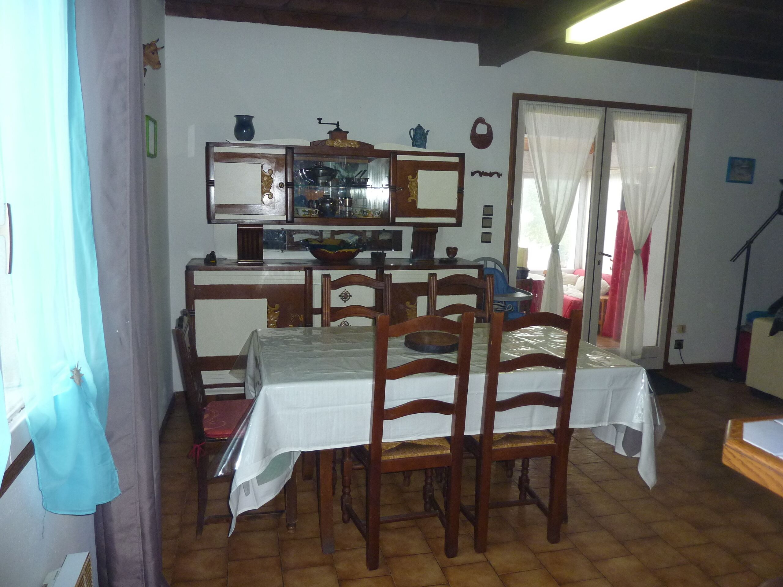 Een eetgedeelte met een tafel voor vier personen, houten stoelen, een keuken met fornuis en kastjes, en een raam met gordijnen.