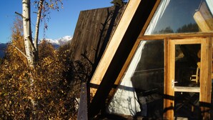 A modern wooden cabin with a slanted roof, large windows, and a view of a snowy mountain range.