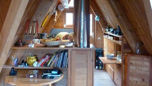 An attic room with wooden beams, a central chimney, a wooden table, shelves with various items, and a window.