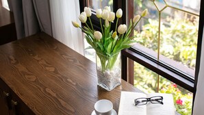 A wooden table with a vase of white tulips, a cup, and an open book with glasses on it, next to a window with a geometric pattern. - Little Charm Hanoi Hostel (Hanoi)