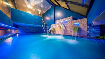 An indoor swimming pool with a waterfall feature, surrounded by stone walls and blue lighting.