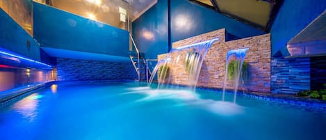An indoor swimming pool with a waterfall feature, surrounded by stone walls and blue lighting.