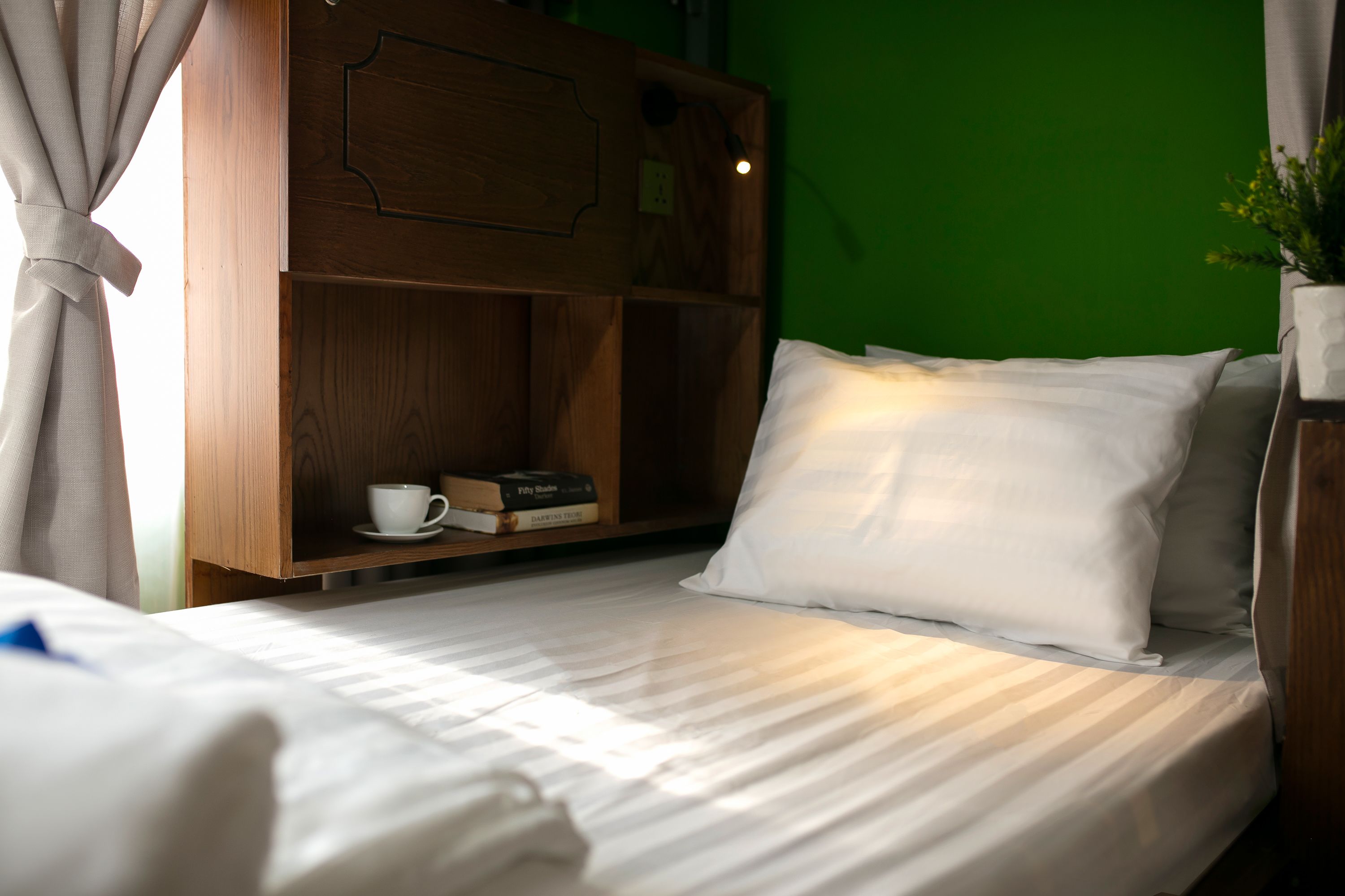 A neatly made bed with white linens, a wooden shelf with books and a cup, and a green wall in the background.
