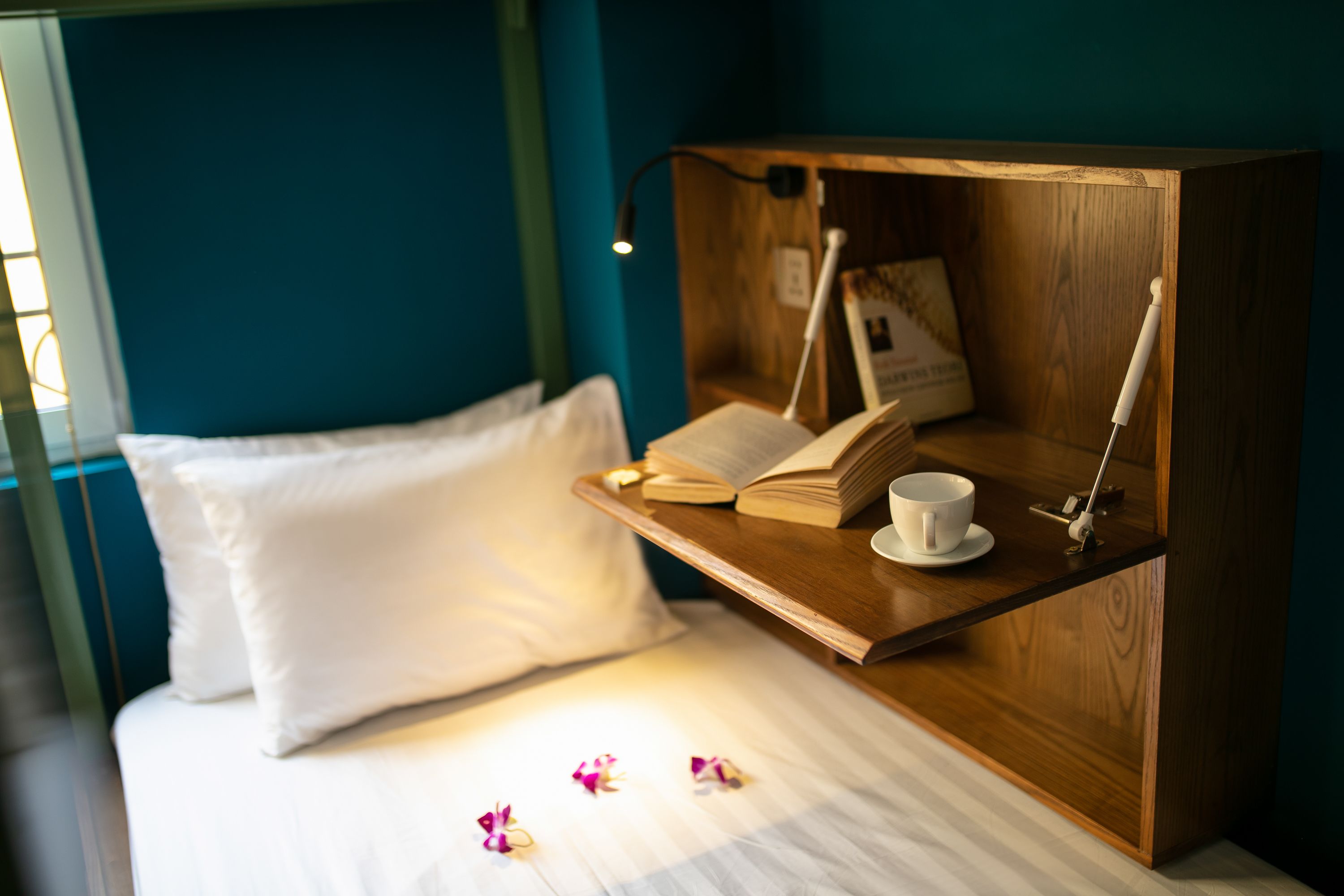 A neatly made bed with white pillows and a wooden bedside table holding a book, a cup, and a lamp.