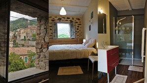 A bedroom with a stone wall, a bed, a window, a bathroom with a shower, and a view of a village.