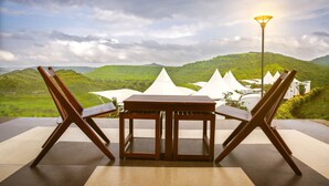 A dining area with a table and chairs, a lamp, and a view of green hills. - Sunny's World (Paud)