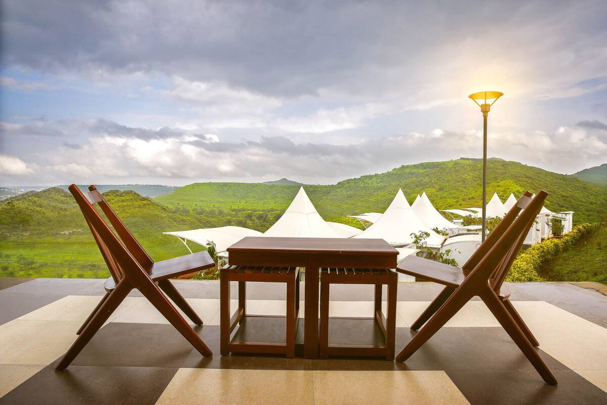 A dining area with a table and chairs, a lamp, and a view of green hills.