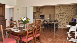 A dining area with a wooden table set for four, surrounded by wooden chairs. A small kitchen is visible in the background with a stone wall.