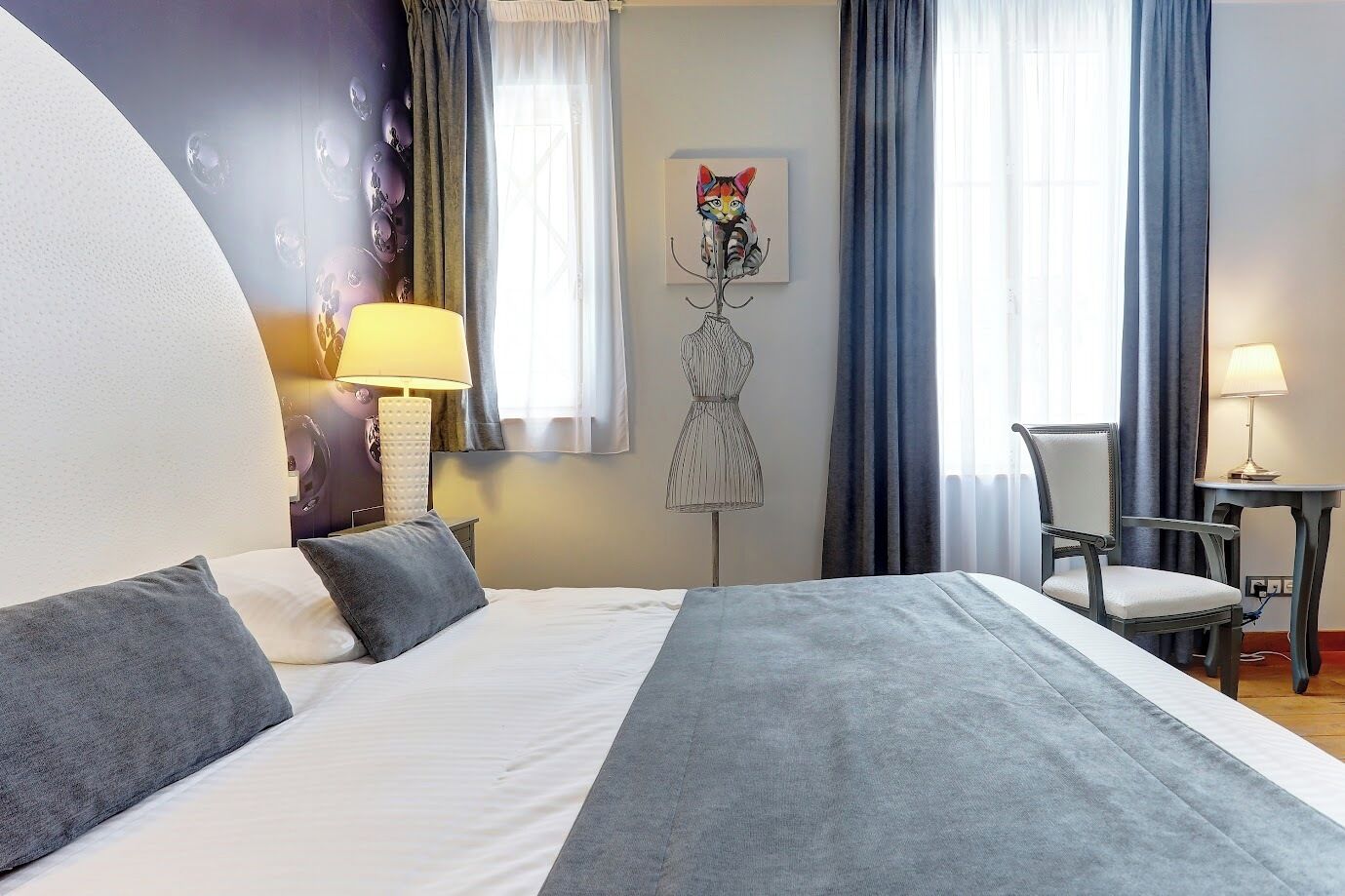 Une chambre d’hôtel avec un lit, une chaise, un bureau, une lampe, un tableau et une fenêtre avec des rideaux.