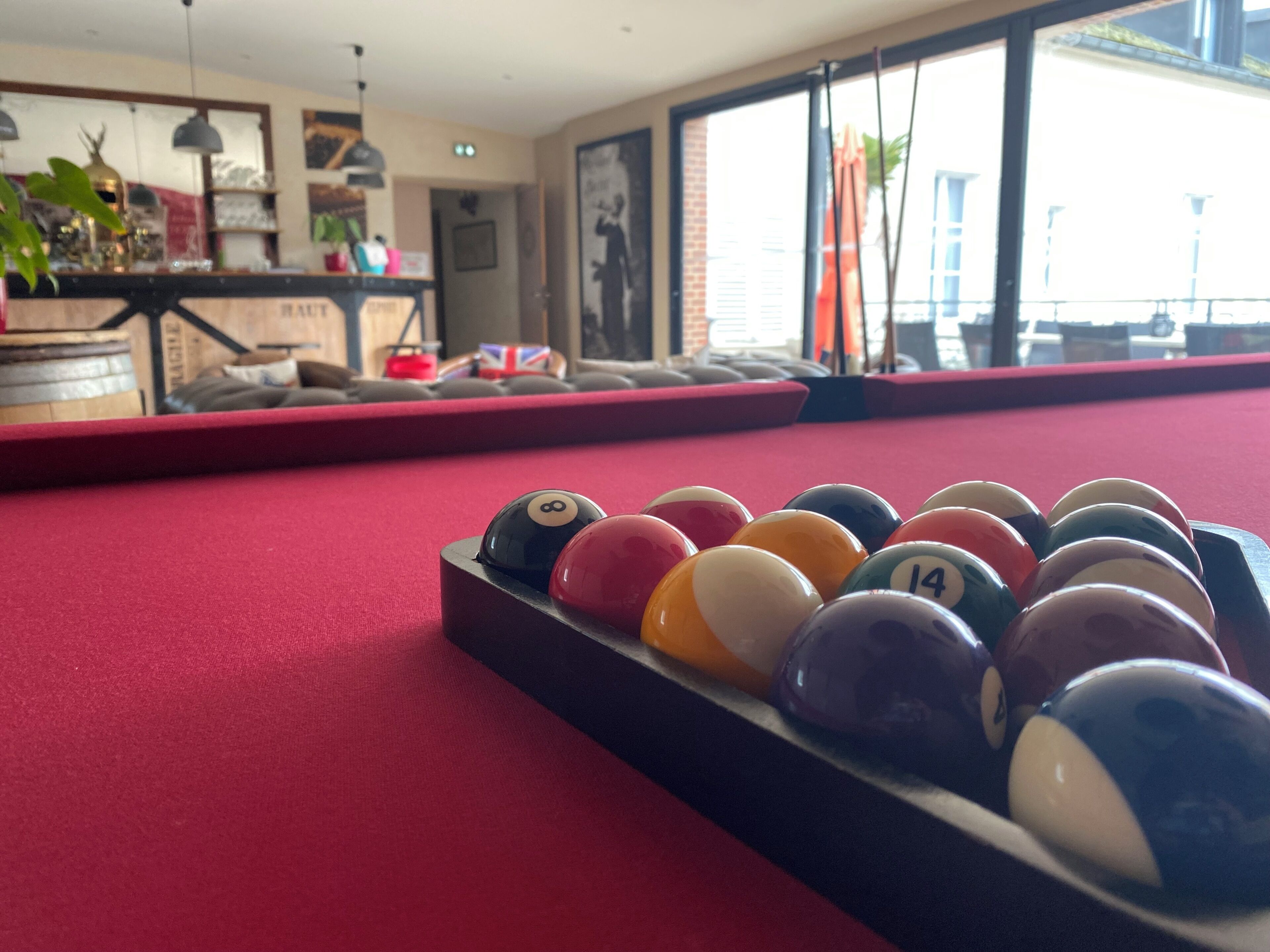 Une table de billard avec des billes numérotées, un drap de jeu rouge et une fenêtre avec des rideaux.