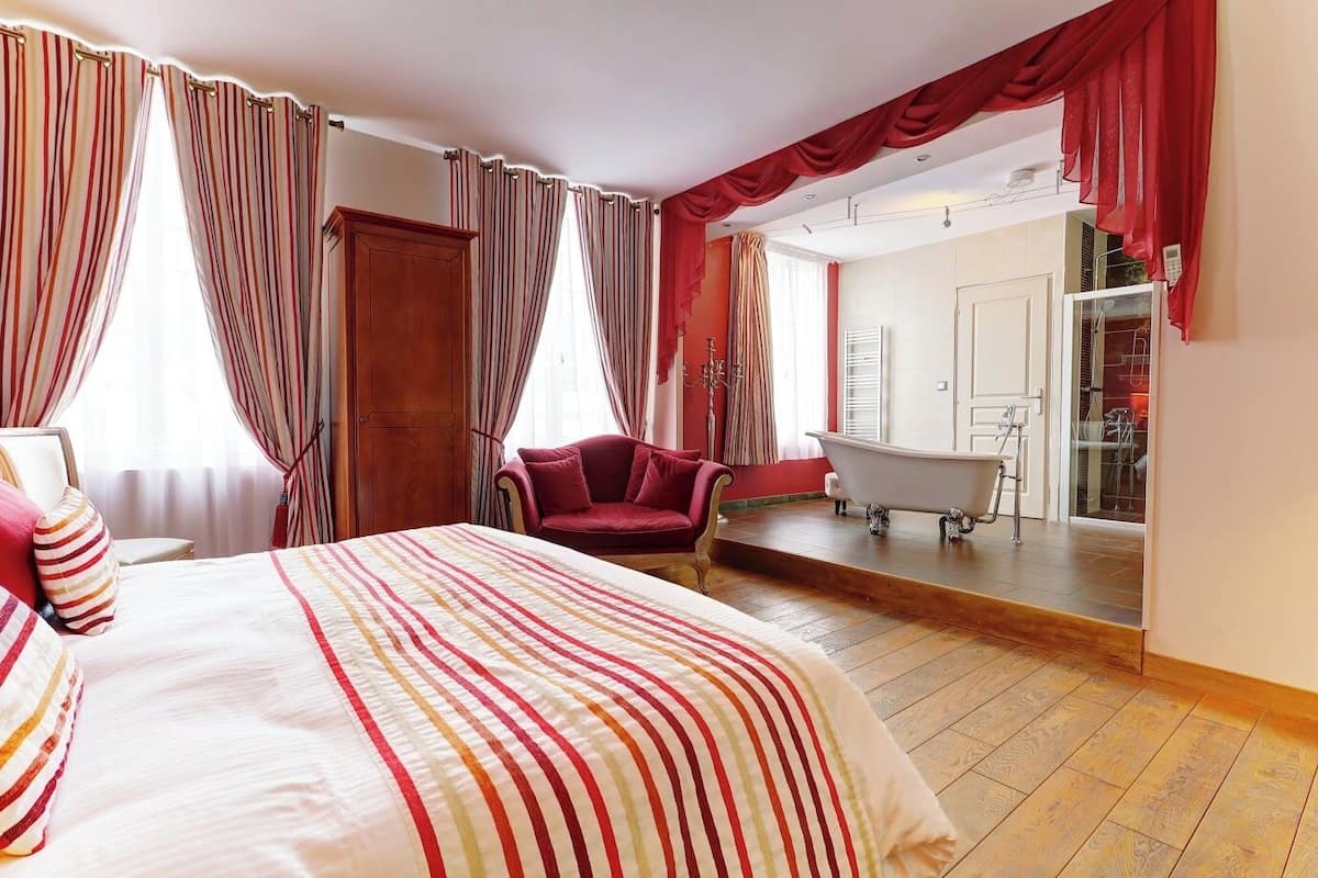 A hotel room with a bed, a red armchair, a clawfoot bathtub, and a large mirror.