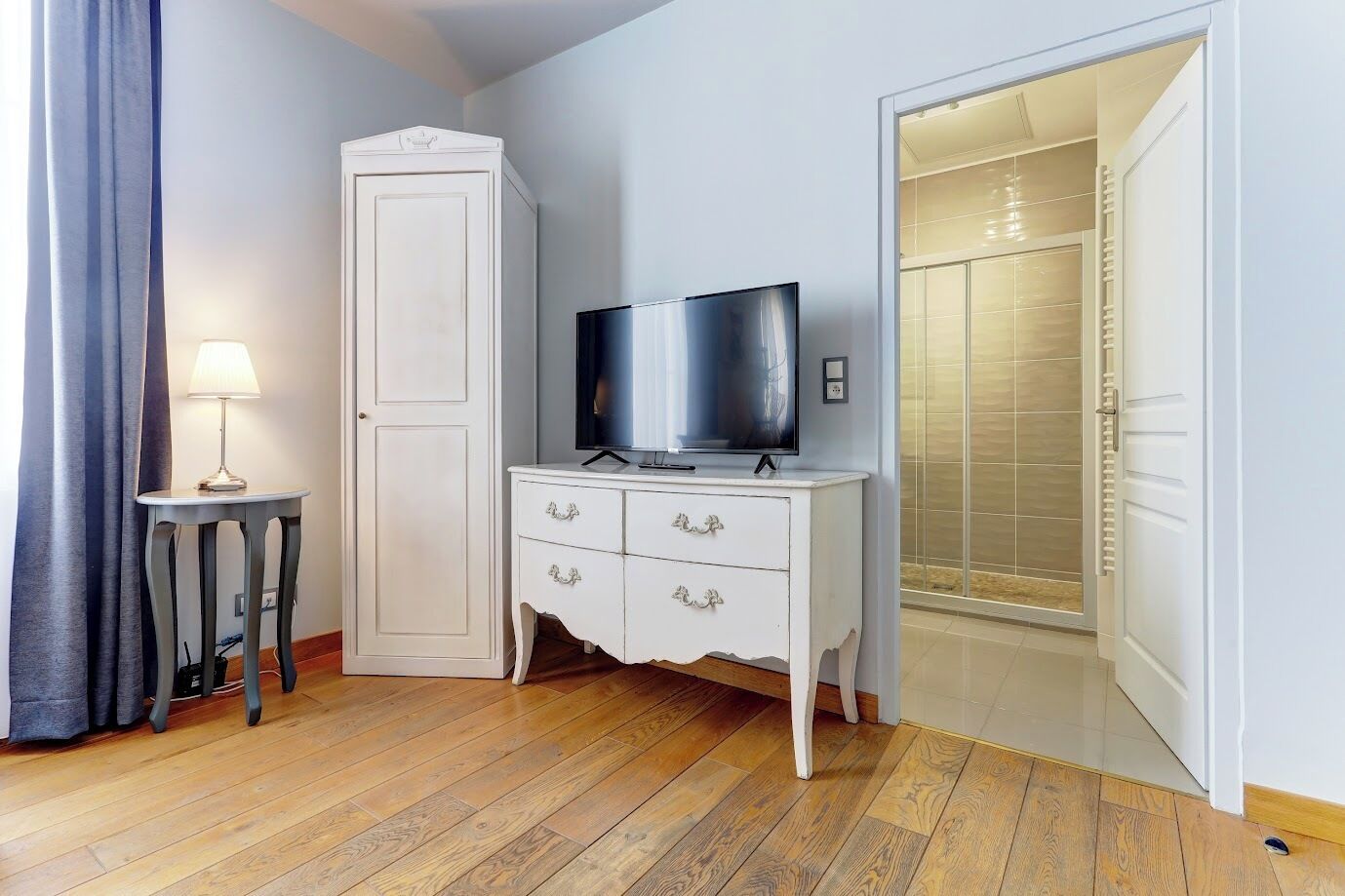 A room with a wooden floor, a white wardrobe, a television on a dresser, a lamp, and a shower area.