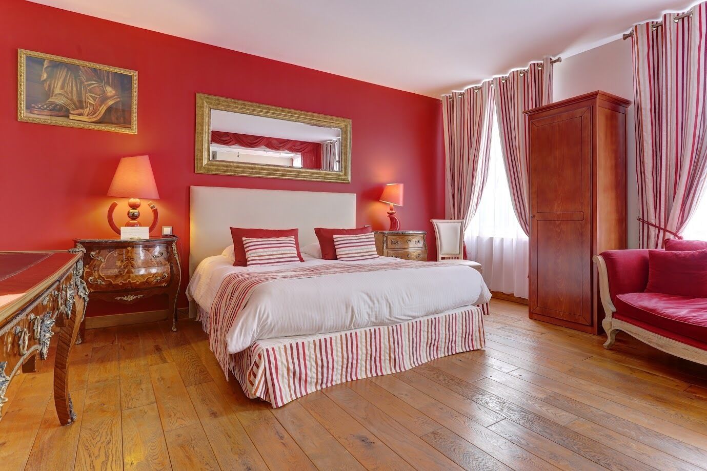 Ein Schlafzimmer mit einem großen Bett, zwei Nachttischen mit Lampen, einem Holzschrank, einem roten Sofa und einem spiegeln an der Wand mit rotem Rahmen.