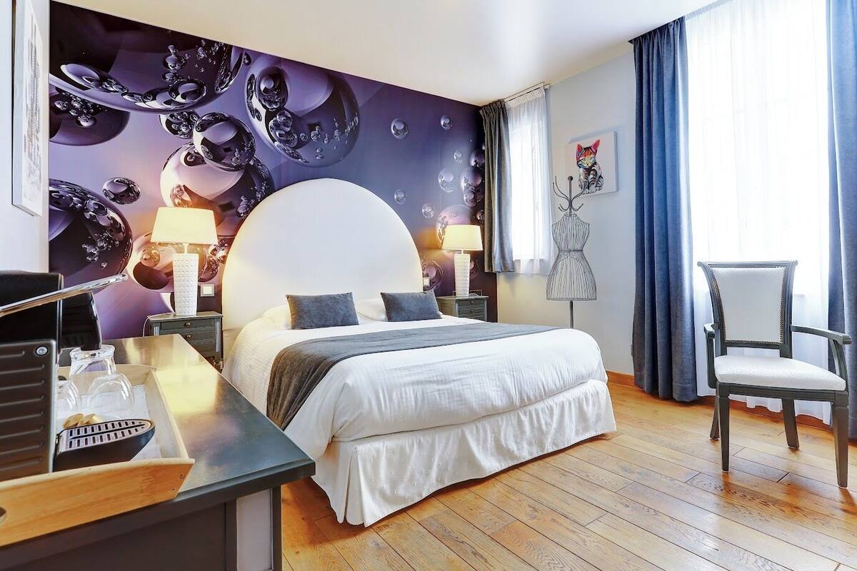 A hotel room with a large bed, bedside tables, a desk with a lamp, a chair, and a wall with a decorative mural.