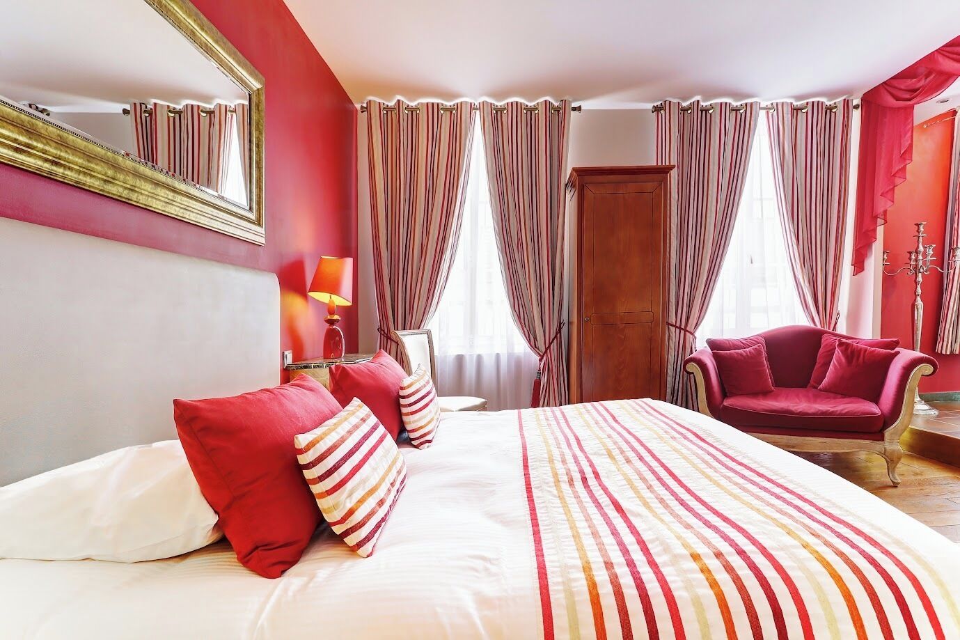 Quarto de hotel com uma cama grande, uma poltrona vermelha, um guarda-roupa de madeira e um espelho na parede.