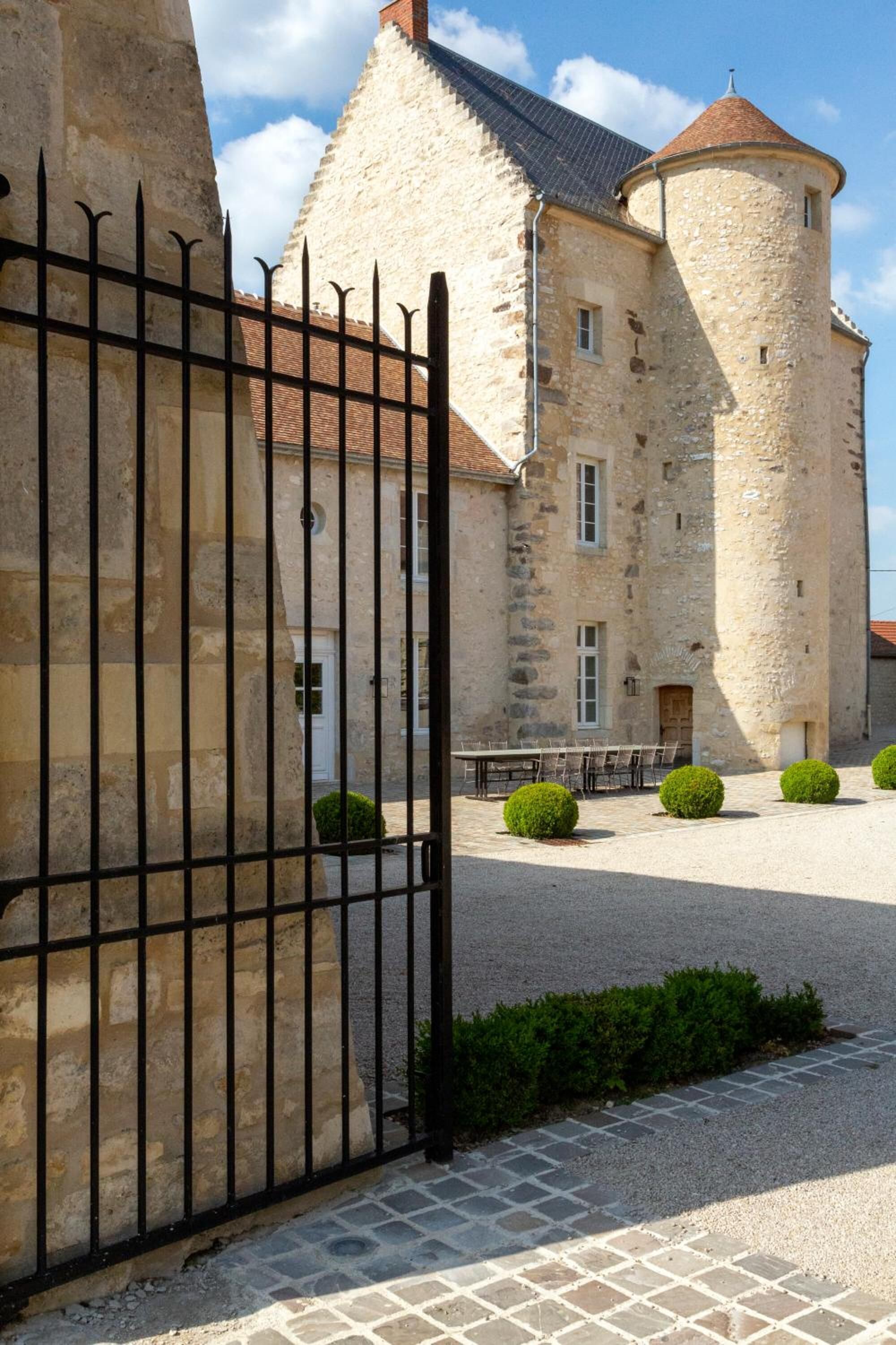 A stone building with a round tower, a gated entrance, and a paved courtyard with trimmed bushes.