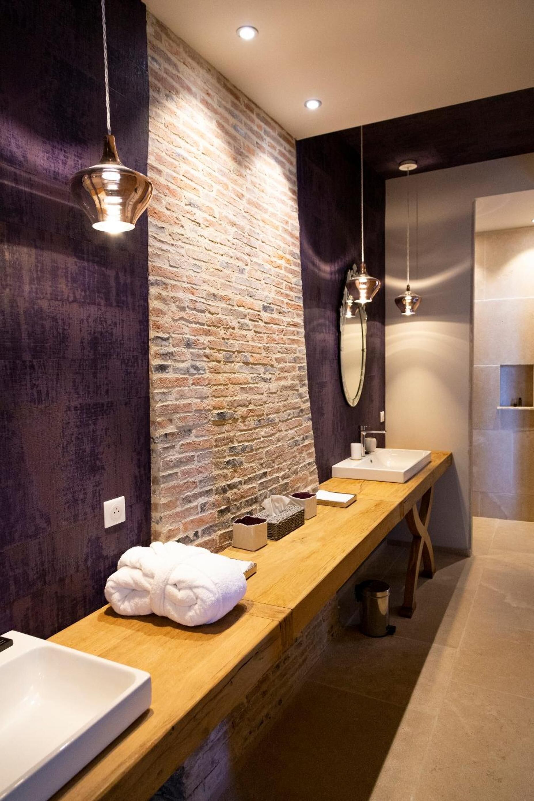 A modern bathroom with a long wooden vanity, a brick accent wall, and pendant lighting.