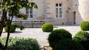A courtyard with stone flooring, a stone building with windows, and neatly trimmed bushes. - La Ferme du Château (Anthenay)