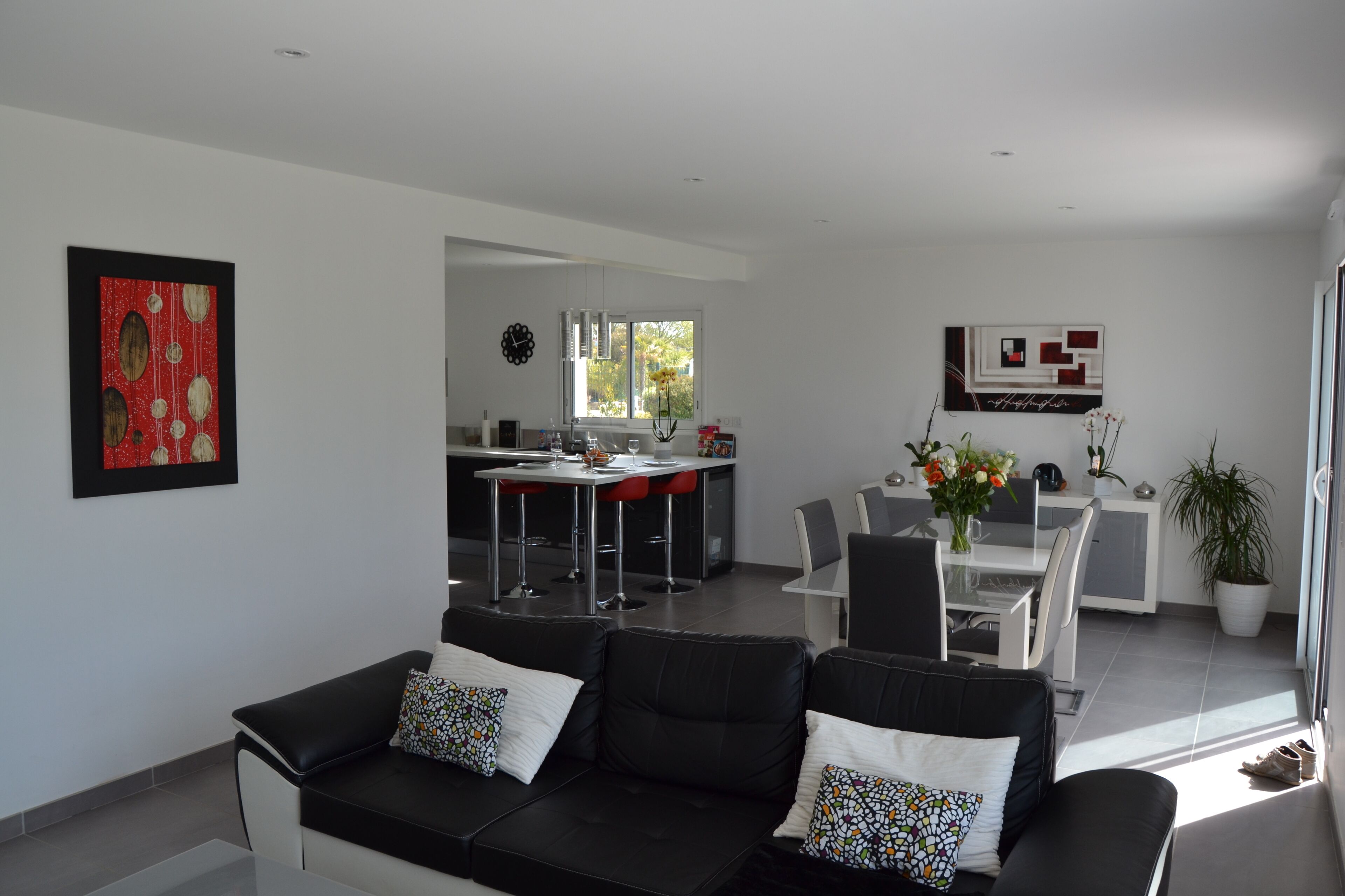 A modern living room with a black leather sofa, a dining area with a white table and chairs, and a kitchen in the background.