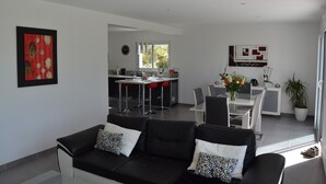 A modern living room with a black leather sofa, a dining area with a white table and chairs, and a kitchen in the background.
