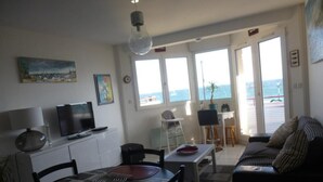 A modern living room with a dining area, a sofa, and a TV. There are large windows with a view of the sea.