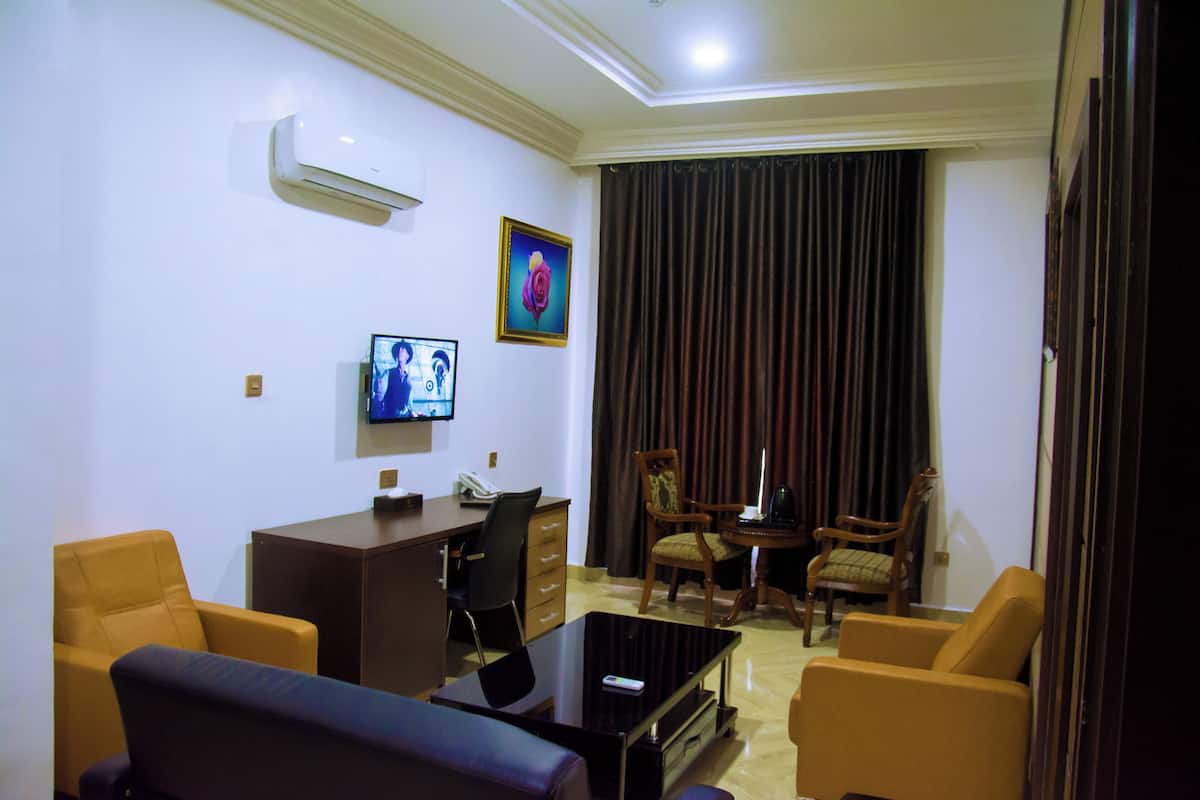 A hotel room with a flat-screen TV, a desk with a telephone, two armchairs, a coffee table, and a large window with curtains.