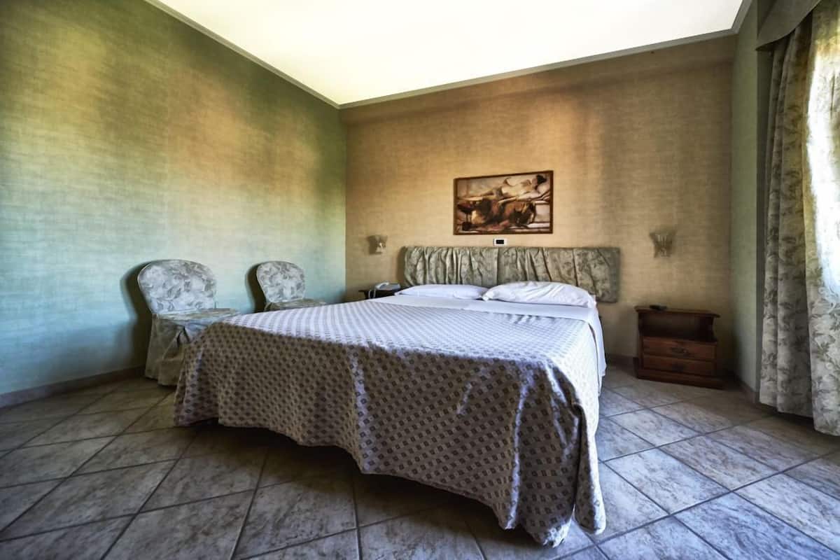 A bedroom with a bed, two chairs, a nightstand, a painting on the wall, and a window with curtains.