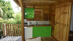 A wooden deck with a wooden chair and a cushion, a kitchen with green cabinets and a wooden door, and a view of greenery outside. - KAZATIPUNCH - CHARMING BUNGALOW IN TROPICAL GARDEN (st francois)