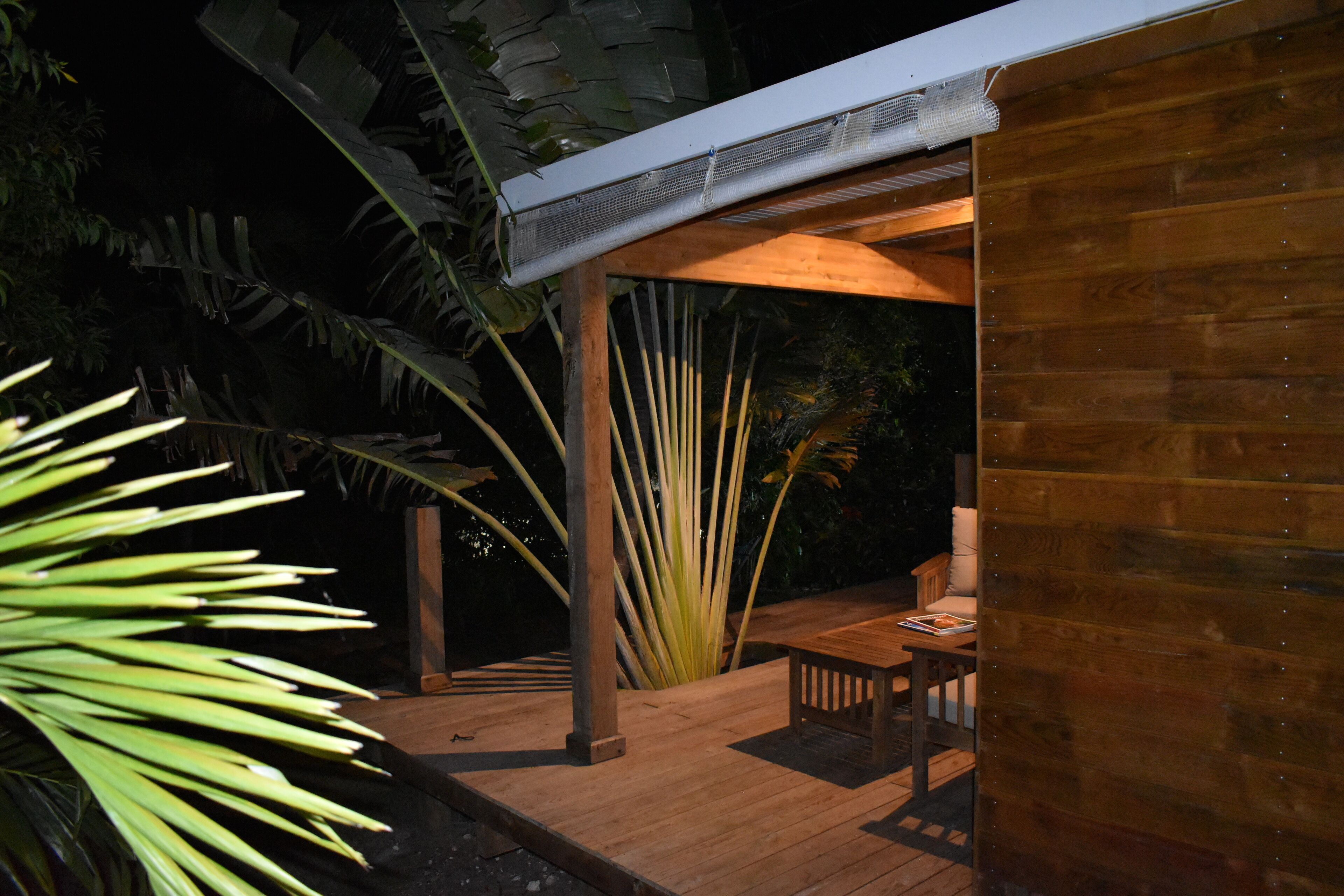 Une terrasse extérieure en bois, dotée d’un espace salon couvert et entourée de plantes tropicales.