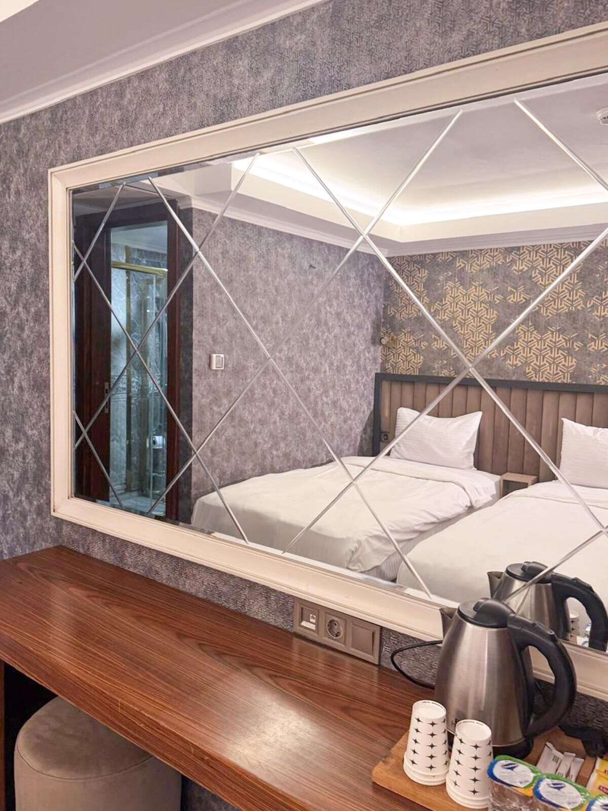 A hotel room with a large mirror, two beds, a wooden desk, a kettle, and a coffee maker.
