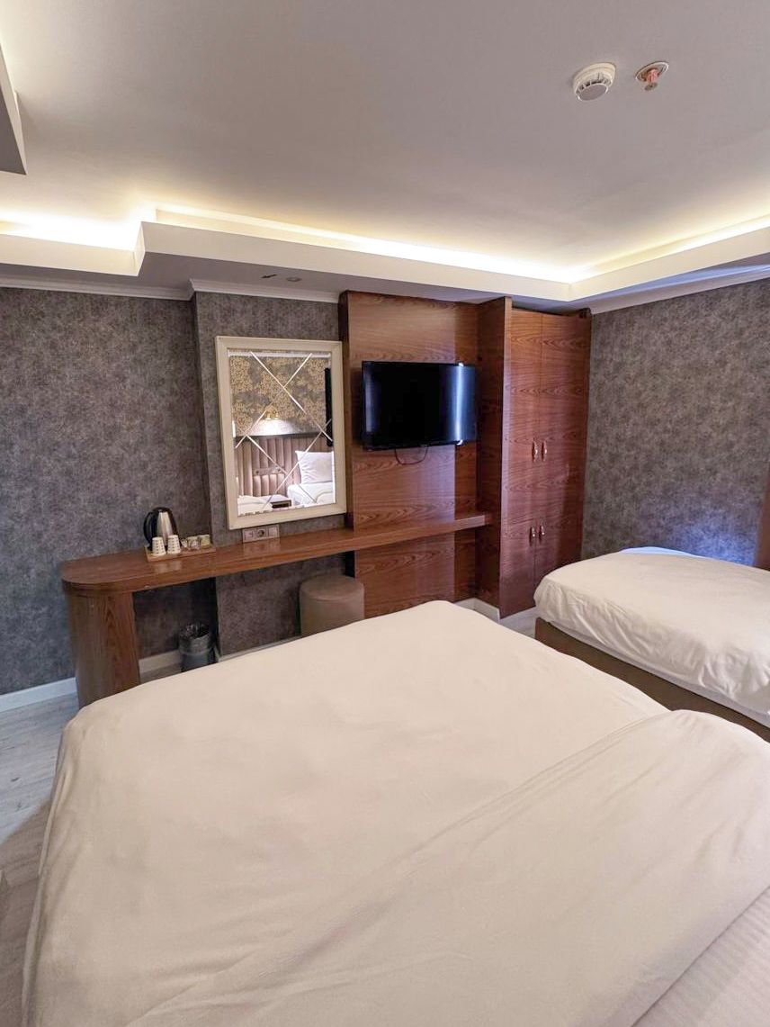 A hotel room with two beds, a TV mounted on the wall, a desk with a mirror, and a small window.