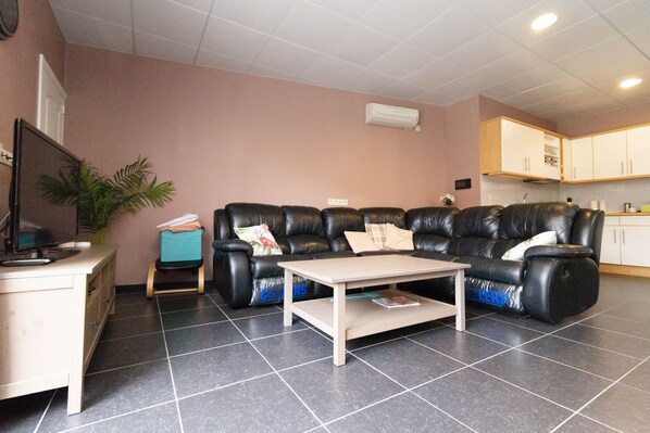 A living room with a black leather sofa, a small coffee table, a desk with a computer, a potted plant, and a kitchenette in the background.