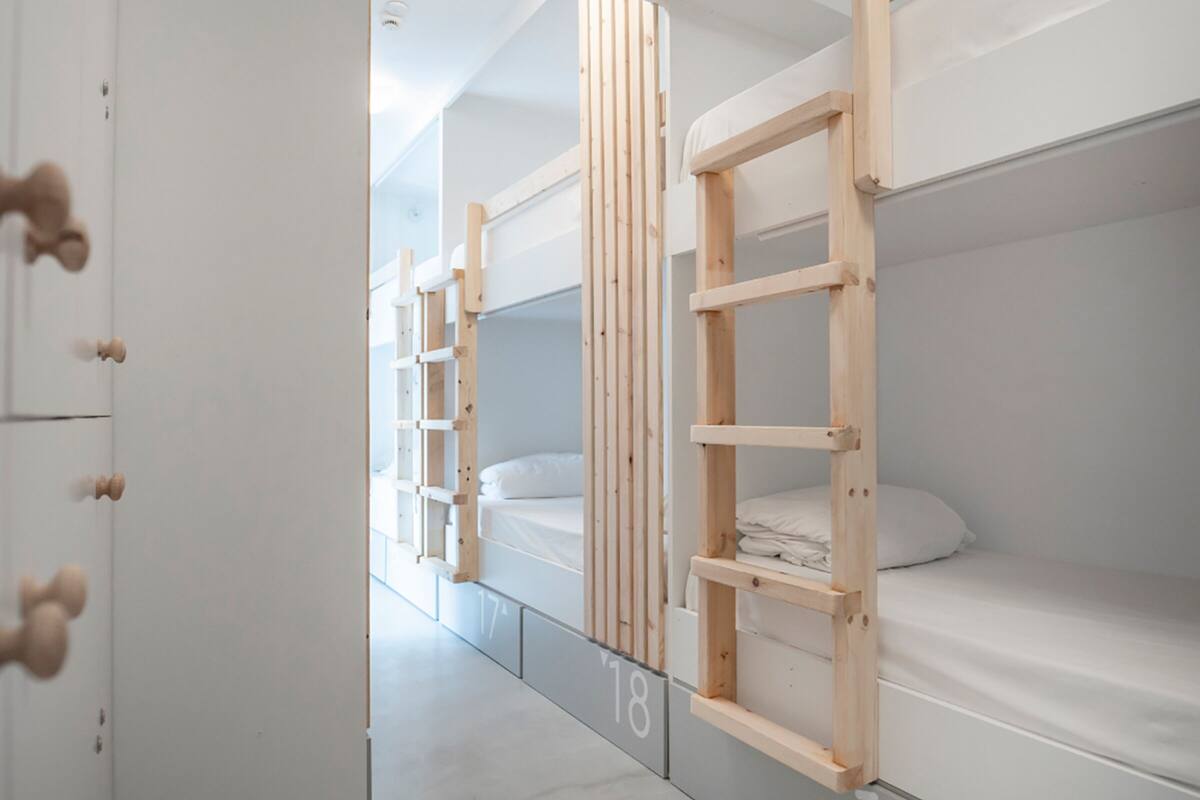 Fila de literas con estructuras de madera y ropa de cama blanca, numeradas 17 y 18, en una habitación moderna y minimalista.