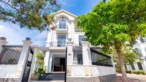 A large, white, two-story building with a gated entrance, a balcony, and a staircase leading to the entrance. - Vihomes Villa (Ho Chi Minh City)