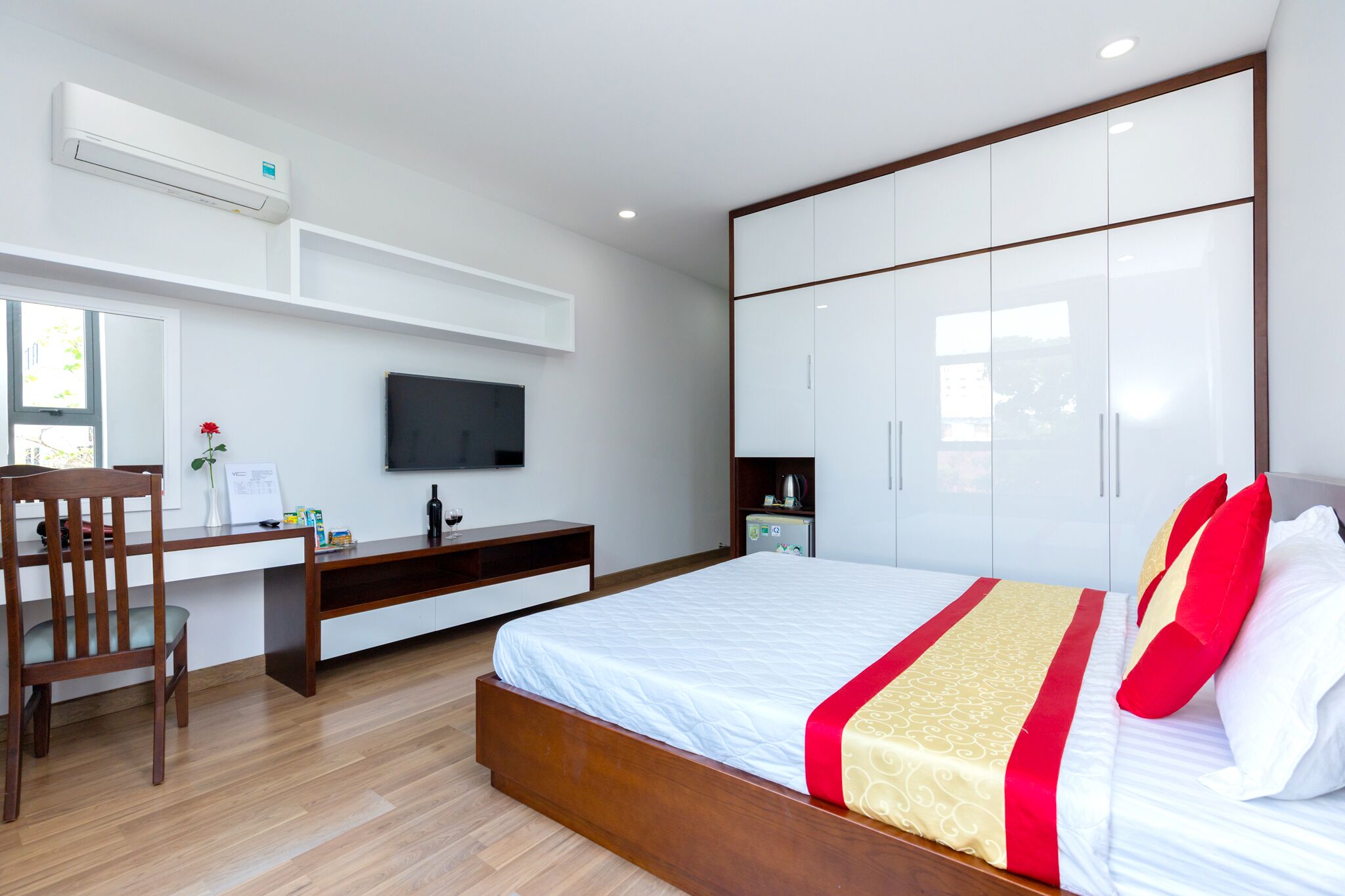 Ein modernes Hotelzimmer mit einem Holztisch, einem Flachbildfernseher, einem Bett mit rot-weißer Bettwäsche und einem Kleiderschrank.