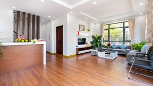 A spacious living area with a wooden floor, a reception desk, a sofa, and a TV on a stand. - Vihomes Villa (Ho Chi Minh City)