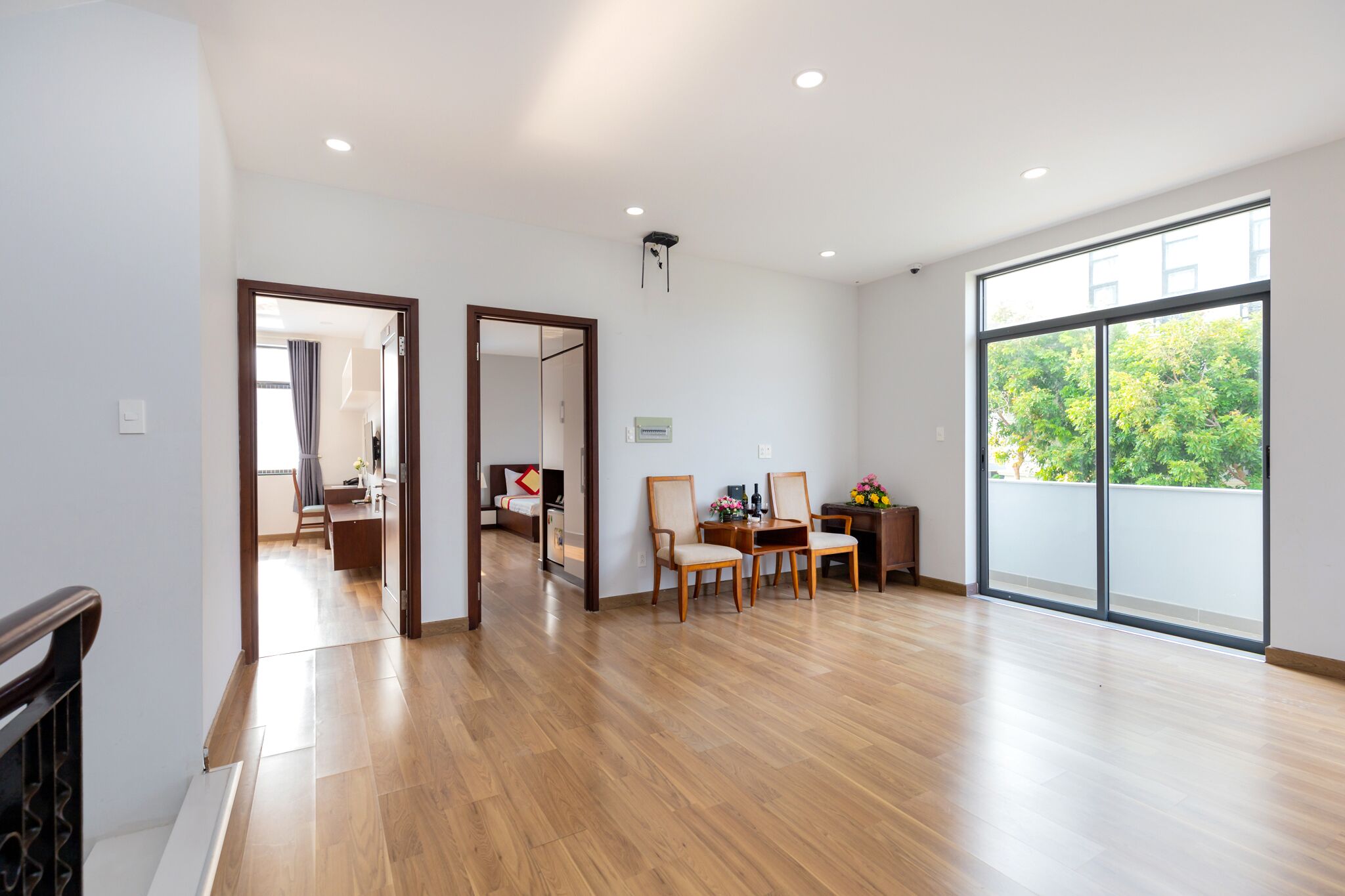 A spacious living area with wooden flooring, a dining table with chairs, a sofa, and a large window offering a view of greenery.