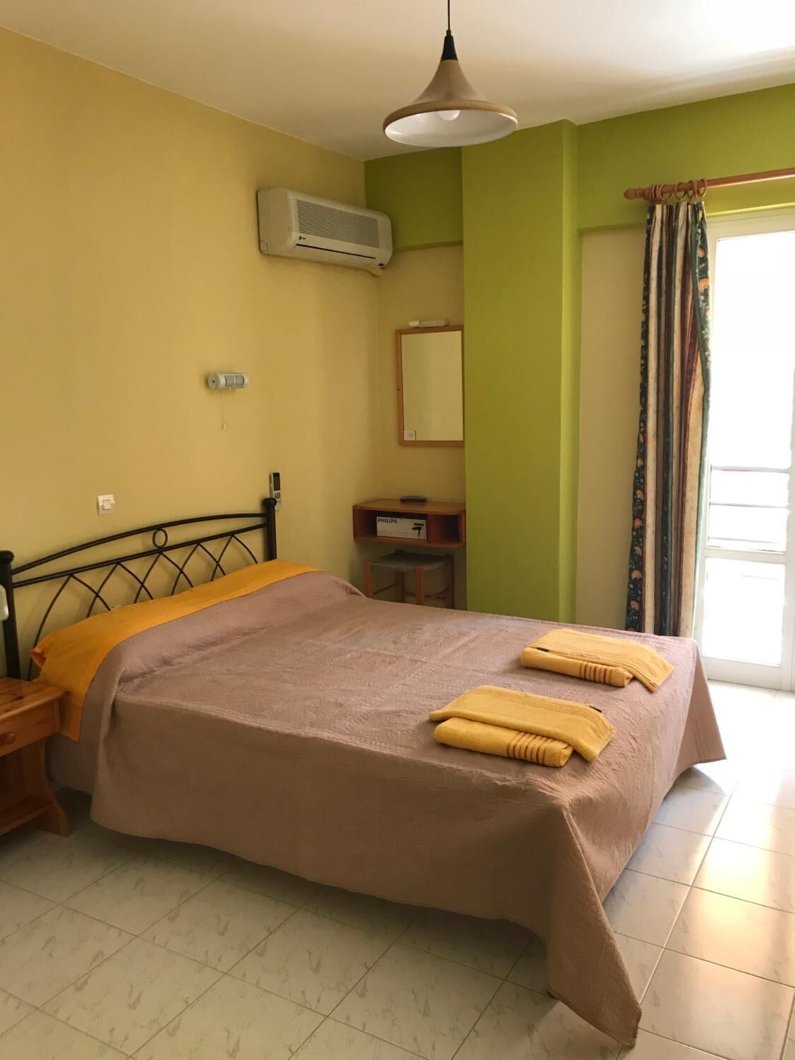 Une chambre d’hôtel comprenant un lit, une table de chevet, une petite table, un appareil de climatisation, un miroir et une fenêtre avec des rideaux.