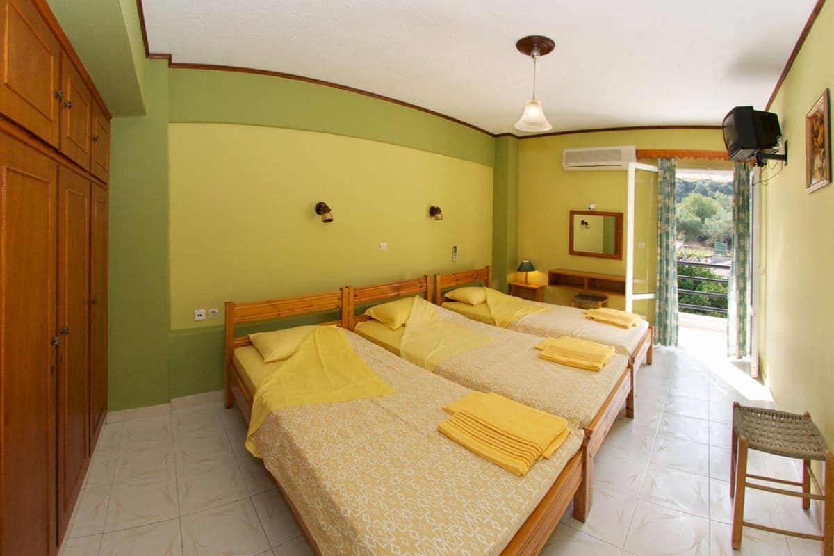 A room with three beds, a wooden wardrobe, a ceiling fan, a mirror, and a window with curtains.