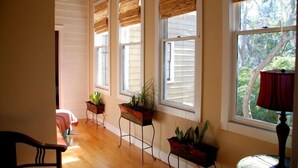 Une pièce avec trois fenêtres, chacune dotée d’un store en bois, un lit recouvert d’une couverture à rayures et deux tables de chevet ornées de plantes en pot.