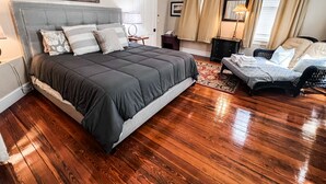 Une chambre à coucher avec un grand lit, une chaise, une table de chevet, une lampe et une fenêtre avec des rideaux.