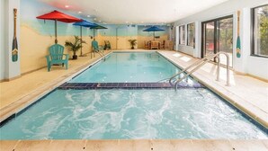 Una piscina interior con superficie de agua cristalina, rodeada de sillas de descanso y sombrillas, y vistas al exterior a través de amplias ventanas.