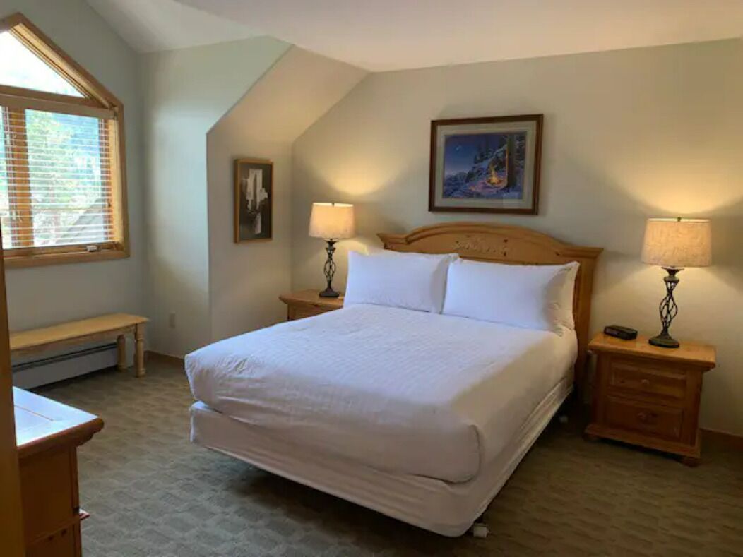 A bedroom with a large bed, two bedside tables with lamps, a bench, a painting on the wall, and a window with blinds.