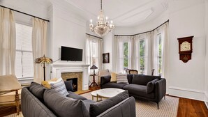A living room with a fireplace, two sofas, a television, and a chandelier. - Newly Renovated 1890 Victorian Downtown Home w/ Heated Pool by Lucky Savannah (Savannah)