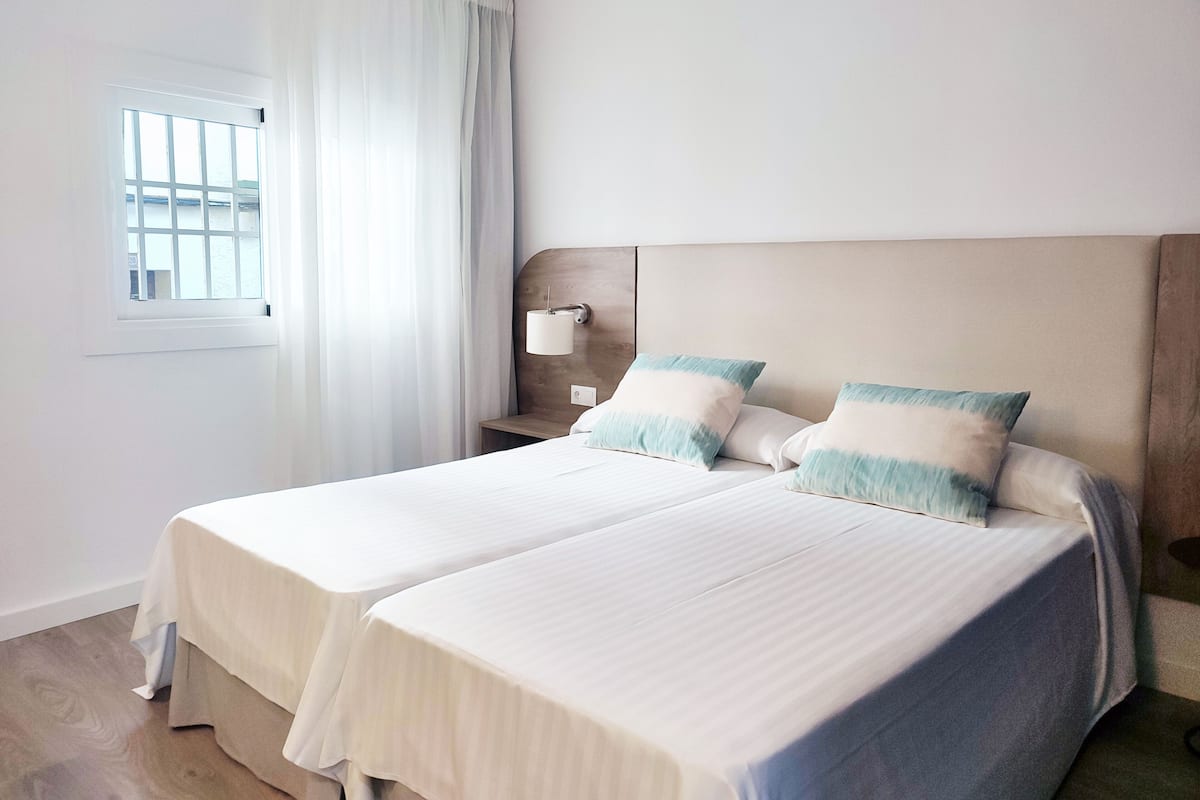 Habitación de hotel con dos camas, una mesita de noche, una lámpara y una ventana con cortinas.
