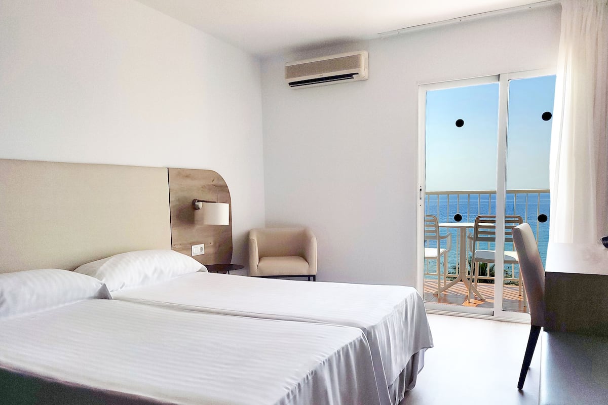 Habitación de hotel con dos camas, una silla, una mesita y un balcón con vista al mar.