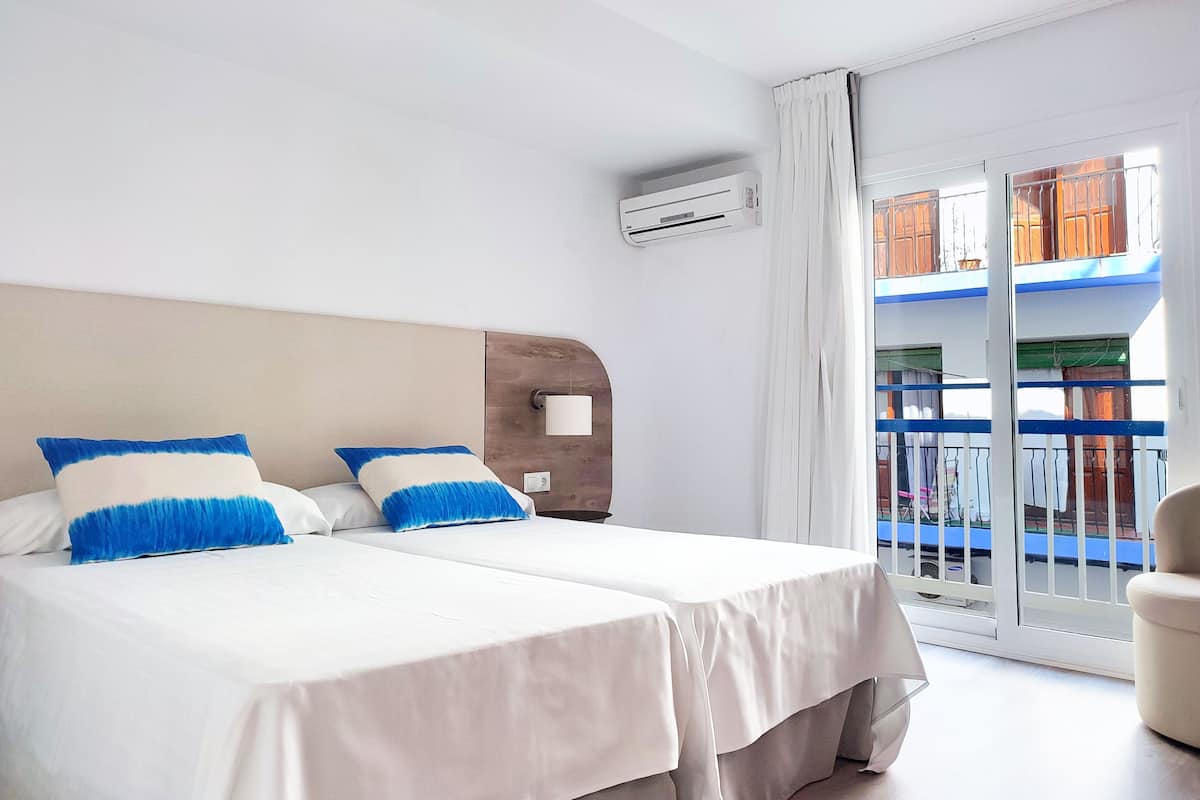 Habitación de hotel con dos camas, un ventanal con vista a un balcón y un equipo de aire acondicionado en la pared.