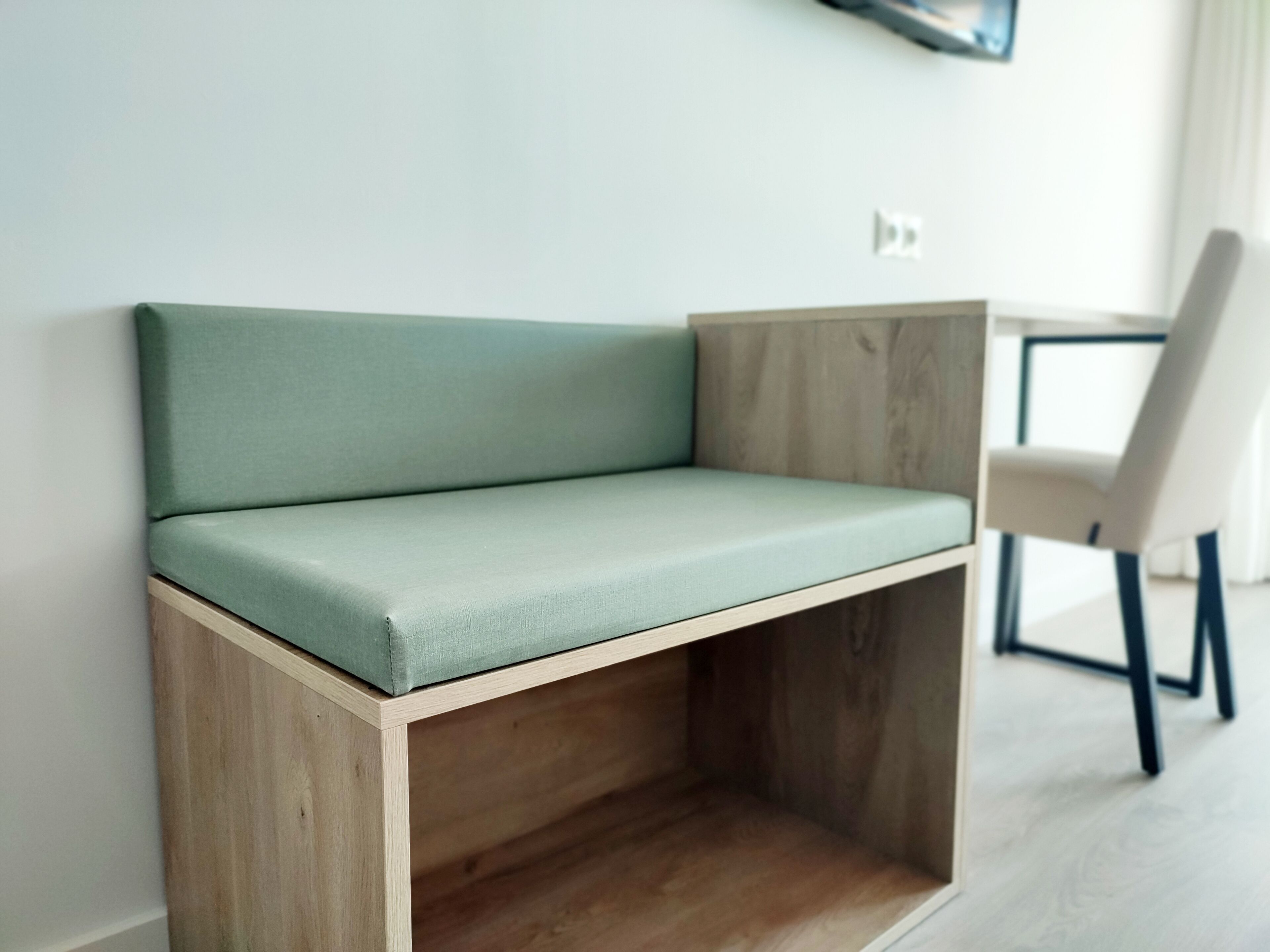 Un canapé d’angle avec une structure en bois et un coussin vert clair, placé contre un mur blanc dans une pièce au plancher en bois, avec une chaise blanche en arrière-plan.