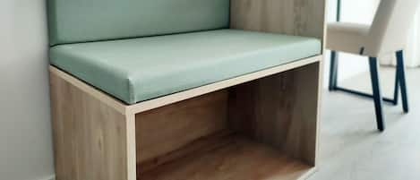 A corner sofa with a wooden frame and a light green cushion, positioned against a white wall in a room with a wooden floor and a white chair in the background.
